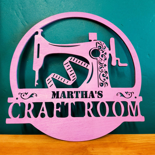 featuring the silhouette of a manual sewing machine with a fabric tape measure below, and space for the owner's name between two scrolls. This Craft Room sign will mark your sanctuary.