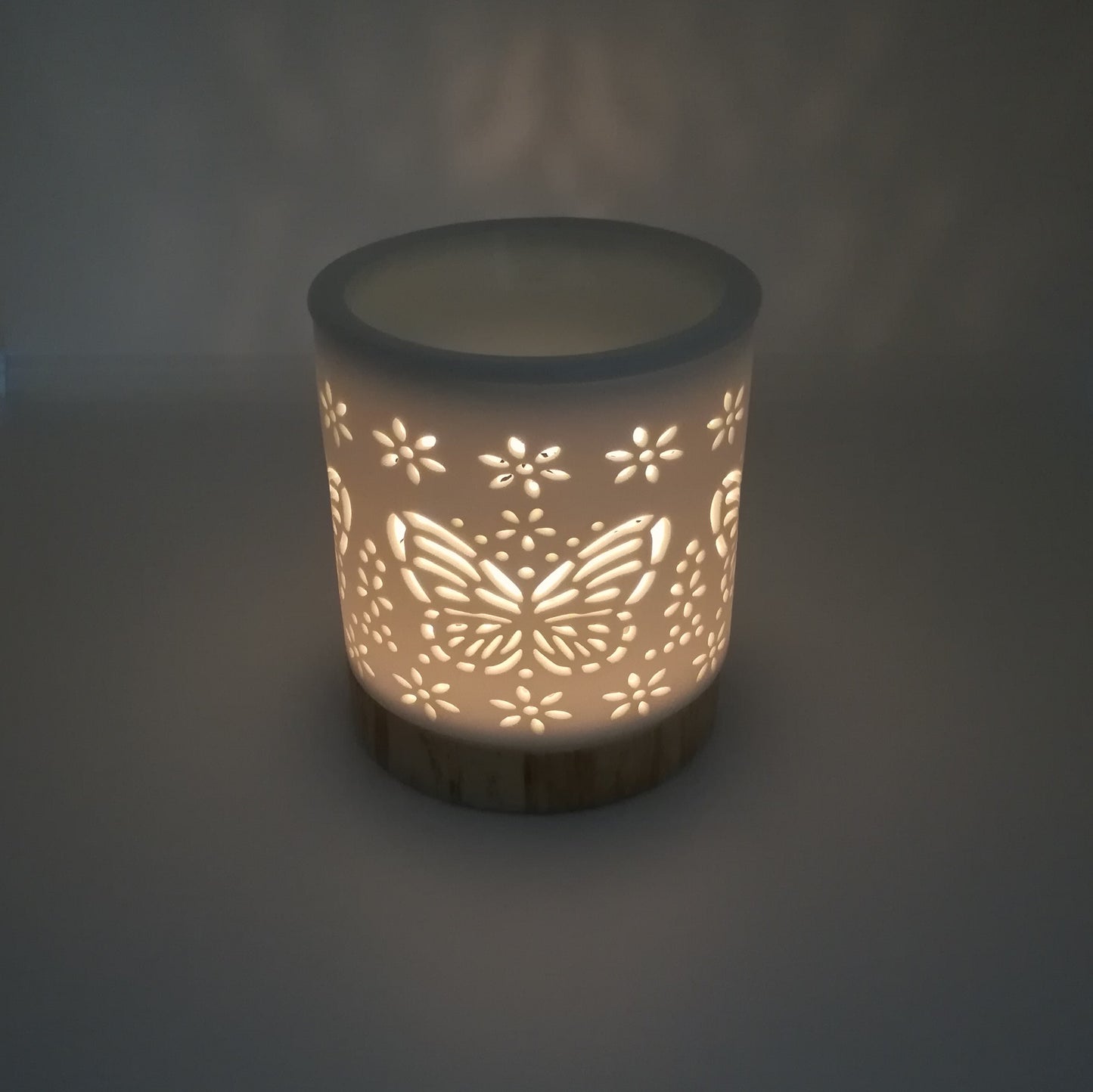 Ceramic white wax melt burner with ceramic wood effect base pictured in semi dark natural light with Butterfly apertures illuminated by the tealight within. (Tealight not included)