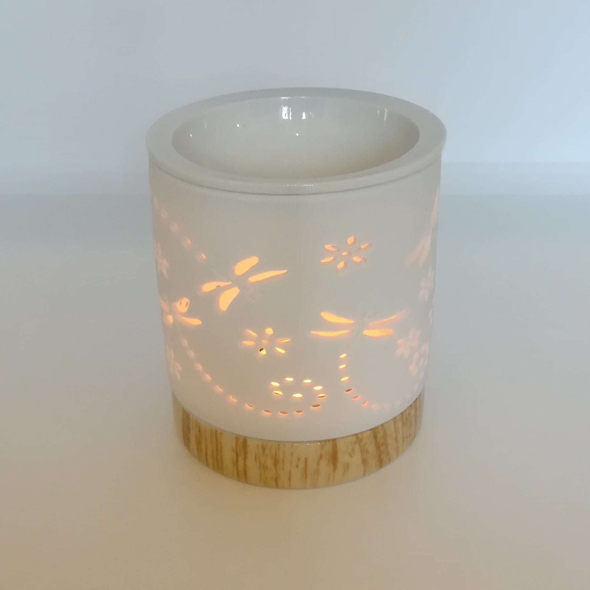 Ceramic white wax melt burner with ceramic wood effect base pictured under natural light with dragonfly apertures illuminated by the tealight within. (Tealight not included).